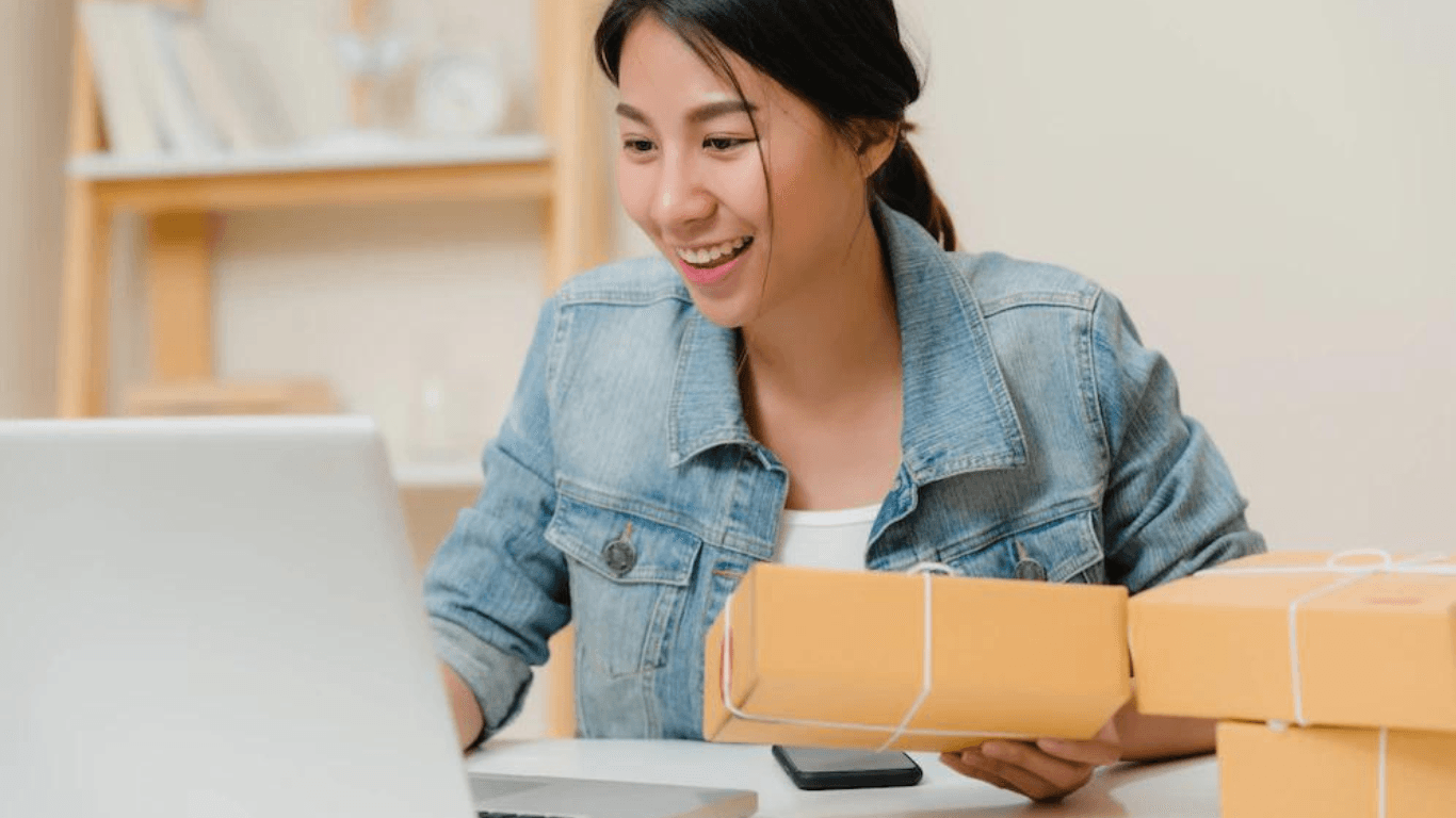 A female online retailer updating her product descriptions on her laptop while holding a parcel